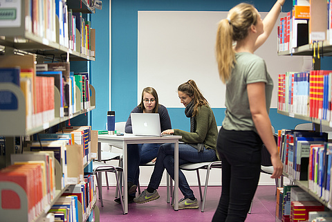 Zwei Personen am Laptop am Tisch, eine Person sucht ein Buch aus einem Regal raus