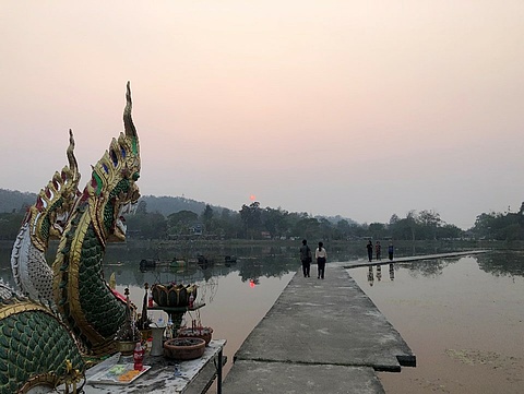 Ein Steg der über einen See geht während eines Sonnenuntergangs. Einige Menschen läufen über des Steg. Links von dem Steg sind zwei Drachenfiguren.