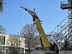 Gelber Lastkran mit Transportseil, im Hintergrund links der Querriegel E-Gebäudes der EHB