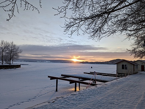 Ein See der zugefrohren ist. Bäume stehen am Rand des Sees. Es leigt Schnee