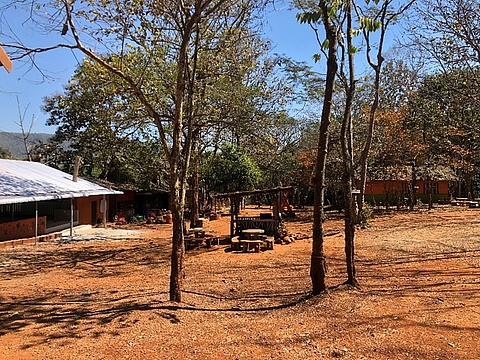 Im Zentrum des Bildes sieht man Pickniktische und Bänke und eine kleine Hütte. Am Rande des Bildes sind zwei Häuser der Boden ist rötlich und auf der offenen Fläche stehen mehrere Bäume